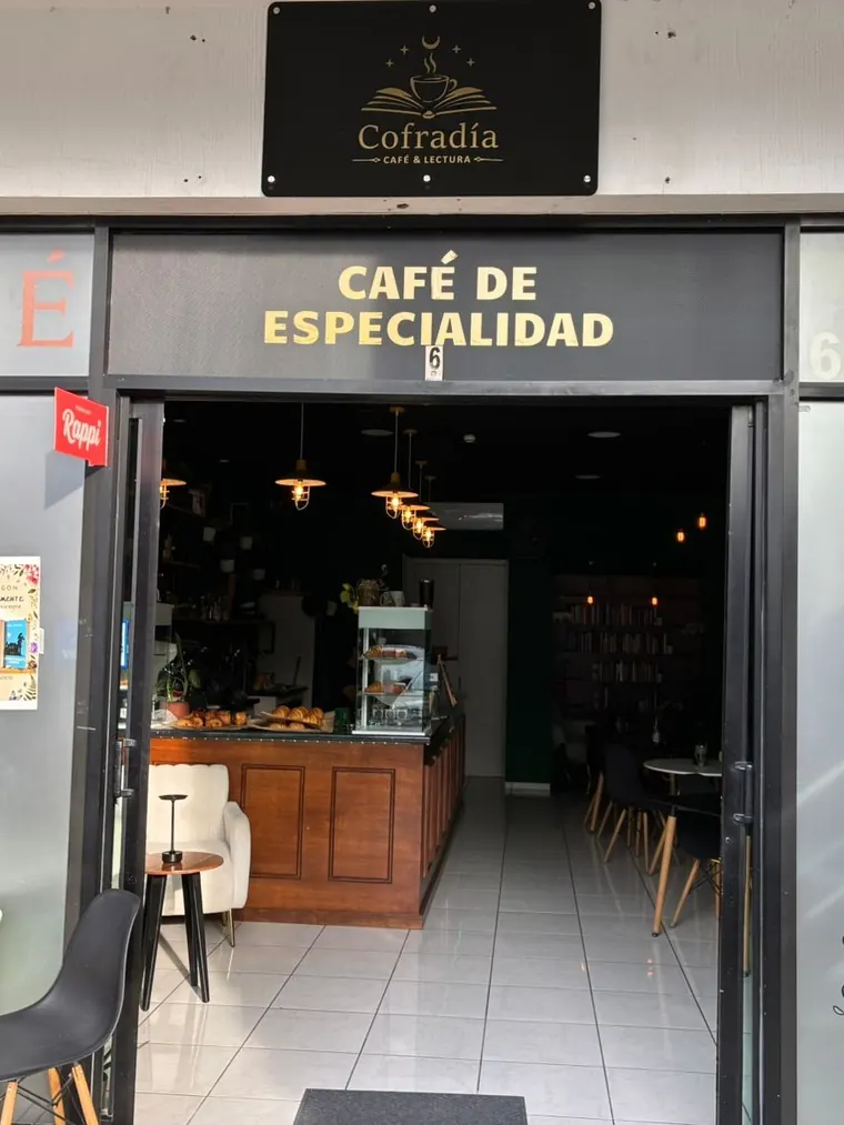 La entrada de Cofradía Café & Lectura desde la calle: el letrero con el logotipo, el rótulo Café de especialidad y la puerta abierta con la barra al fondo